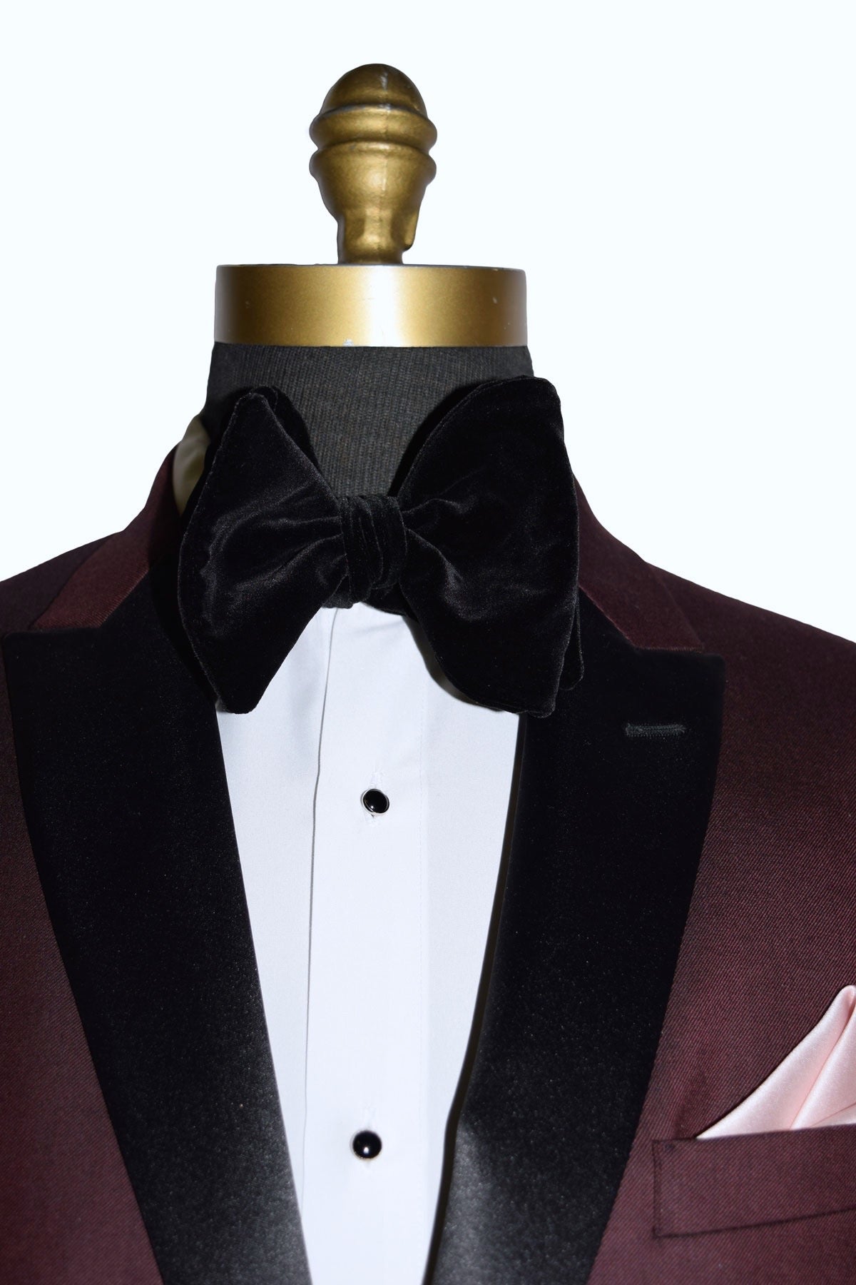 close up front view of burgundy tuxedo with black peak lapels shown with white tuxedo shirt and large black velvet bowtie on a mannequin against a white background