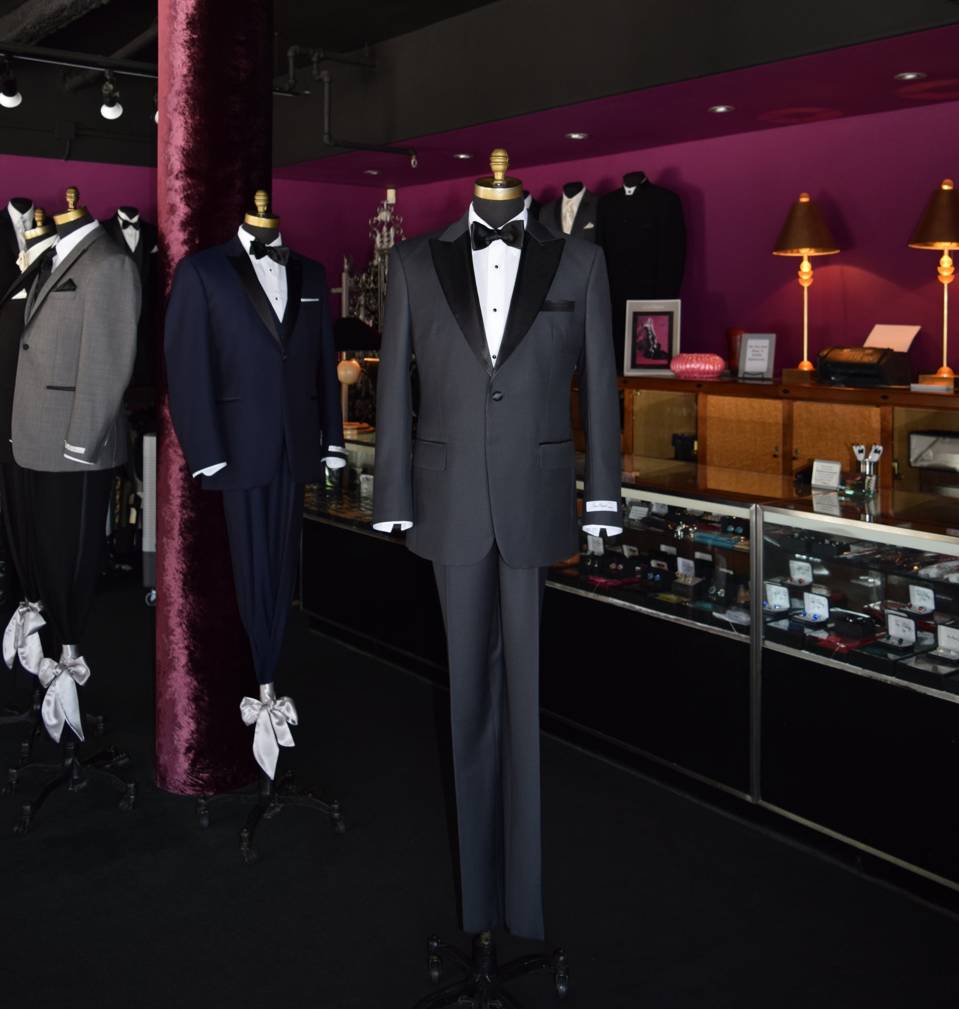 Mannequins wearing a charcoal peak lapel in a tuxedo shop setting with purple walls and display cases.