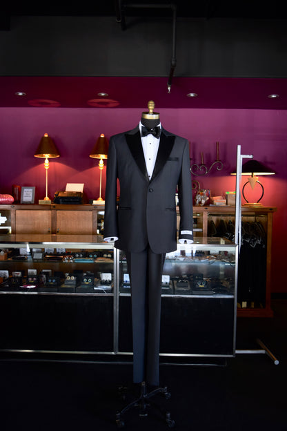 charcoal peak lapel tuxedo wearing a white shirt and black bowtie on a mannequin against a dark background with decorative elements.