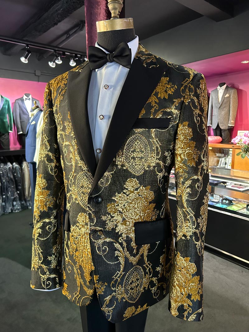 close up front view of mannequin wearing a gold and black patterned tuxedo with black satin bowtie and white tuxedo shirt with pearl studs and cufflinks inside a tuxedo shop