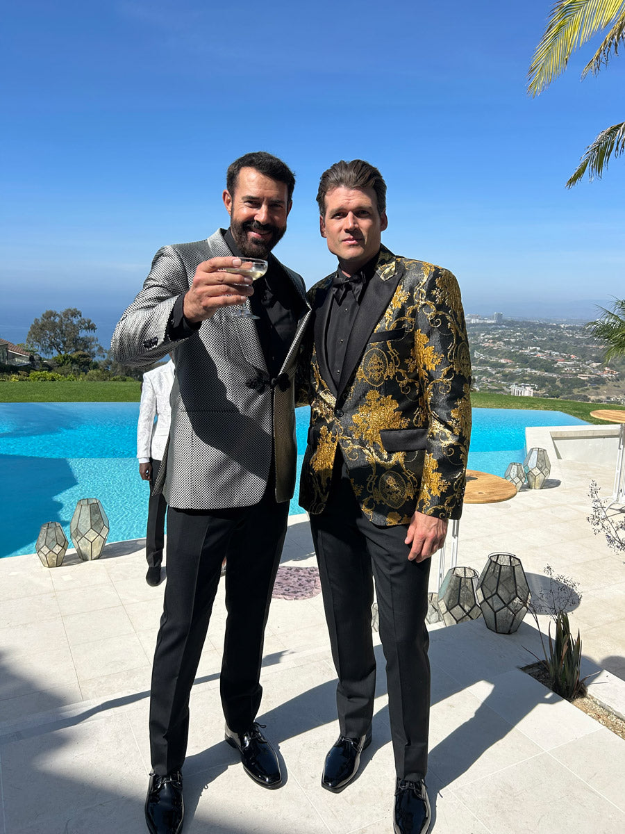 Two men in formal attire standing by a pool with a scenic background and one man is wearing a gold brocade tuxedo with black pants