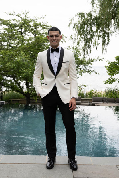 Younger hispanic man wearing an ivory tuxedo with black shawl collar standing by a pool with trees.