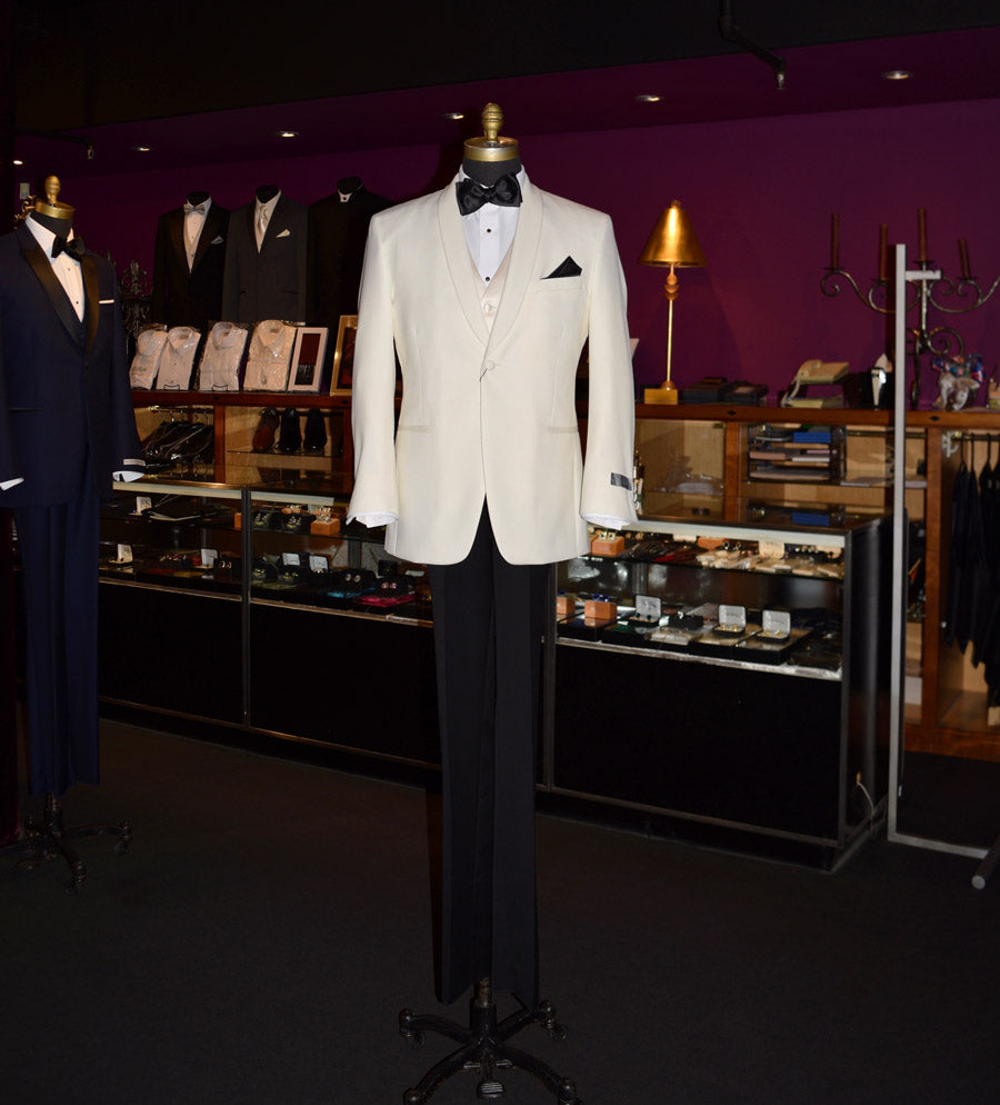 An ivory dinner jacket on a mannequin with black tuxedo pants with a white shirt and an ivory satin vest and black bowtie inside a fancy tuxedo shop