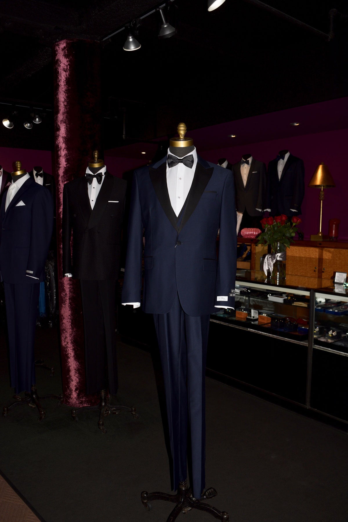 life-size mannequin Navy blue tuxedo with black lapels on a mannequin wearing a black bow tie inside of a fine mens store with other tuxedos against the wall