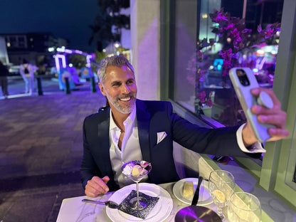Handsome man wearing a navy blue tuxedo with blue lapel taking a selfie at an outdoor dinner table with a dessert and drink.