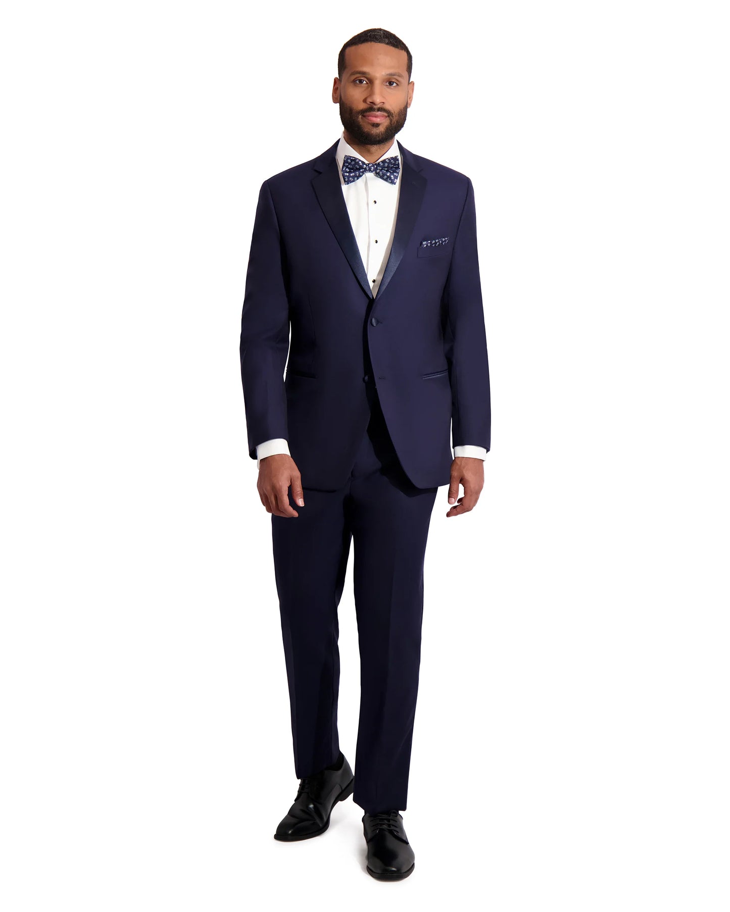 Man wearing a navy blue notch lapel tuxedo by with a blue polkadot bowtie against a white background. 