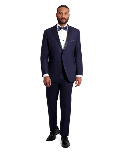 Man wearing a navy blue notch lapel tuxedo by with a blue polkadot bowtie against a white background. 
