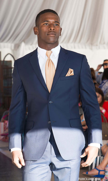 Man wearing a navy slim-fit suit with a light gold tie and pocket square, standing indoors with audience in background