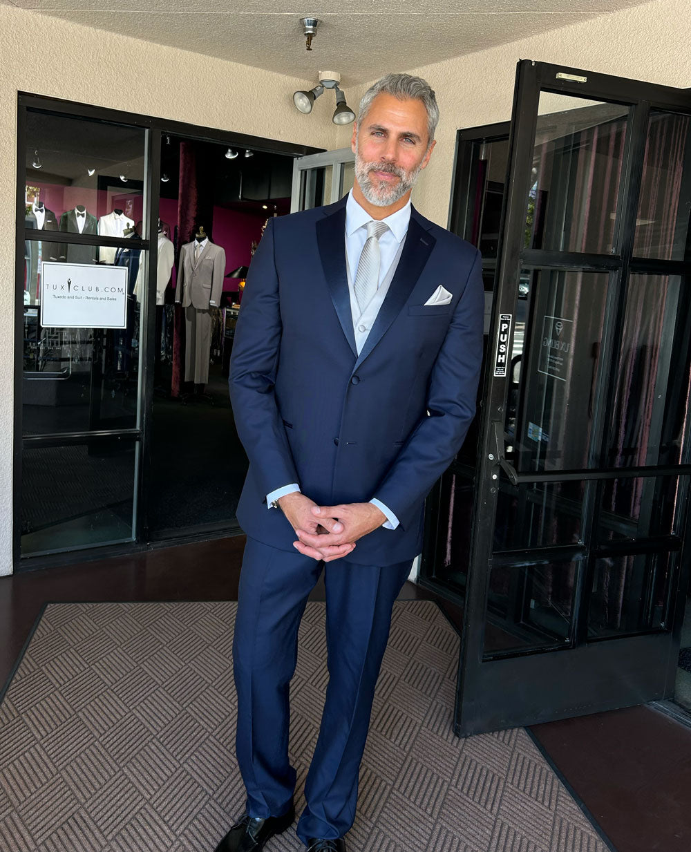 Man in a navy blue tuxedo standing in a tuxedo shop with mannequins in the background