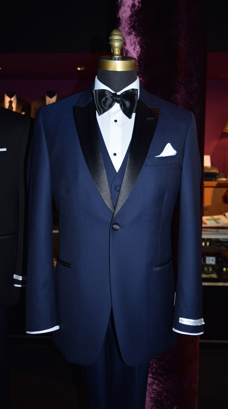 mannequin wearing a 3 piece navy blue tuxedo with black peal lapels with matching navy tuxedo pants with white shirt and black satin bowtie inside a high-end tuxedo shop with other tuxedos on display