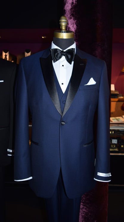 mannequin wearing a 3 piece navy blue tuxedo with black peal lapels with matching navy tuxedo pants with white shirt and black satin bowtie inside a high-end tuxedo shop with other tuxedos on display