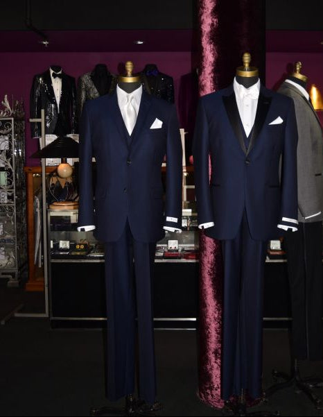 two mannequins wearing a navy blue tuxedo one with a white long dress tie and the other one with a white satin bowtie inside a high end tuxedo shop