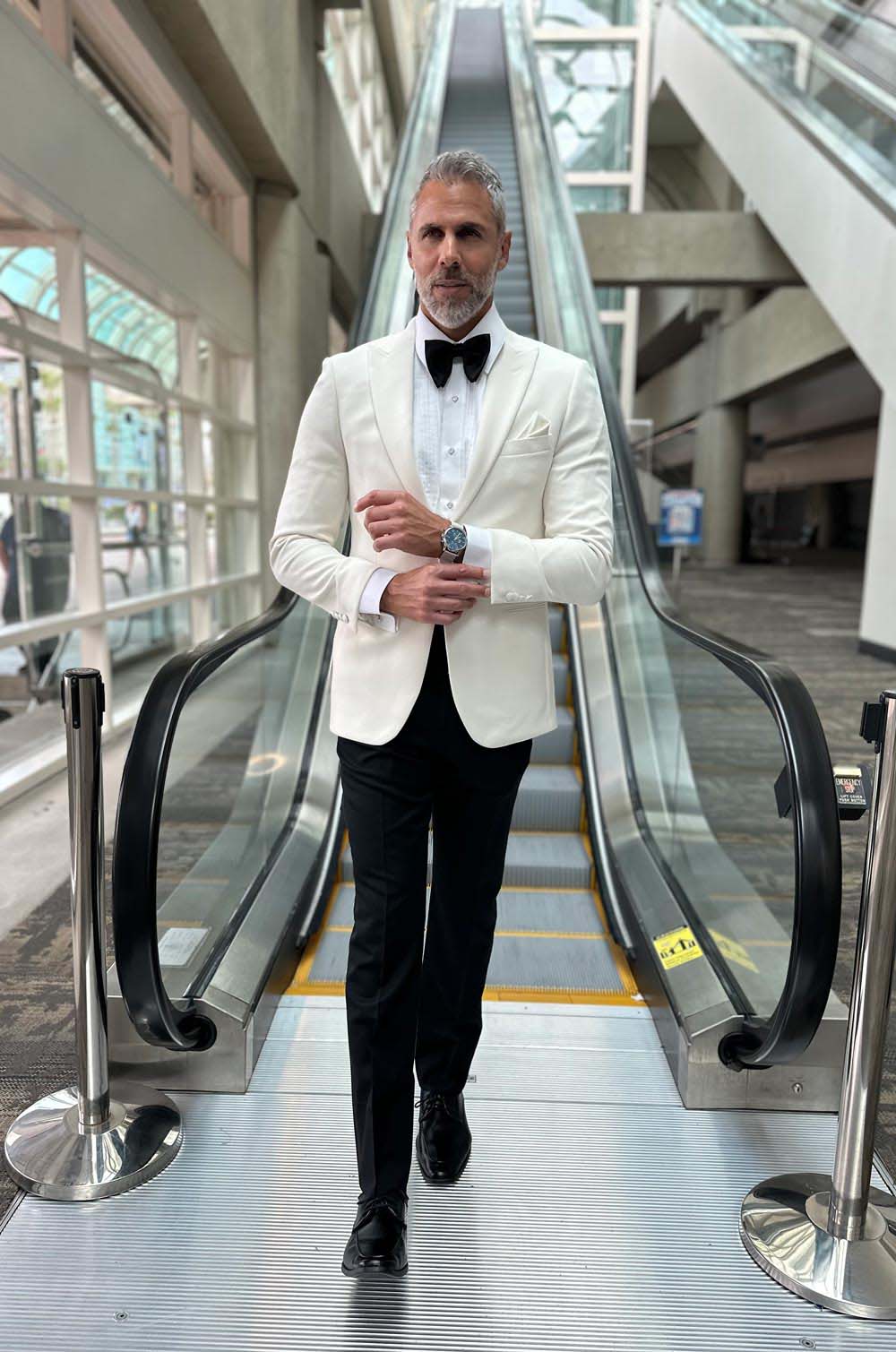 handsome man wearing an ivory peak lapel dinner jacket with a white tuxedo shirt and black bowtie coming down an escalator 