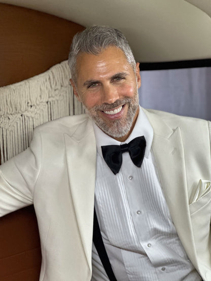Handsome man wearing  off-white peak lapel tuxedo from tuxclub, with black satin bowtie and black suspenders while sitting in a vintage car.