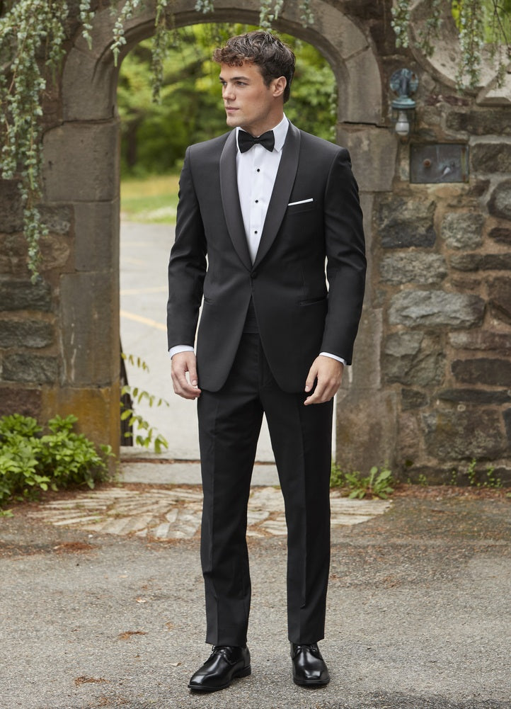 Handsome nan in a black shawl collar tuxedo standing in front of a stone archway.