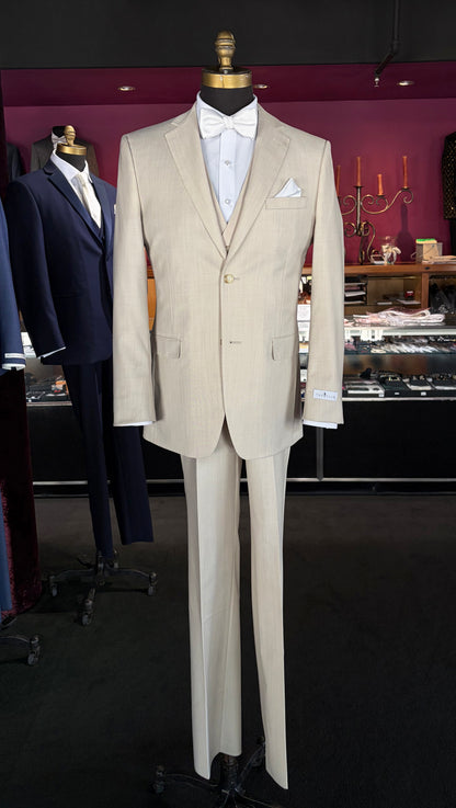 A tan fitted two-piece suit displayed on a mannequin torso, styled with a white dress shirt, white bow tie, and white pocket square, set against a dark boutique interior.