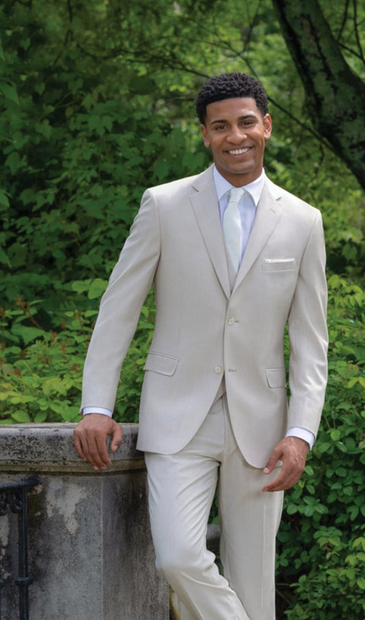 A man wearing a tan three-piece suit with a white dress shirt and white necktie, standing outdoors against a background of green foliage.