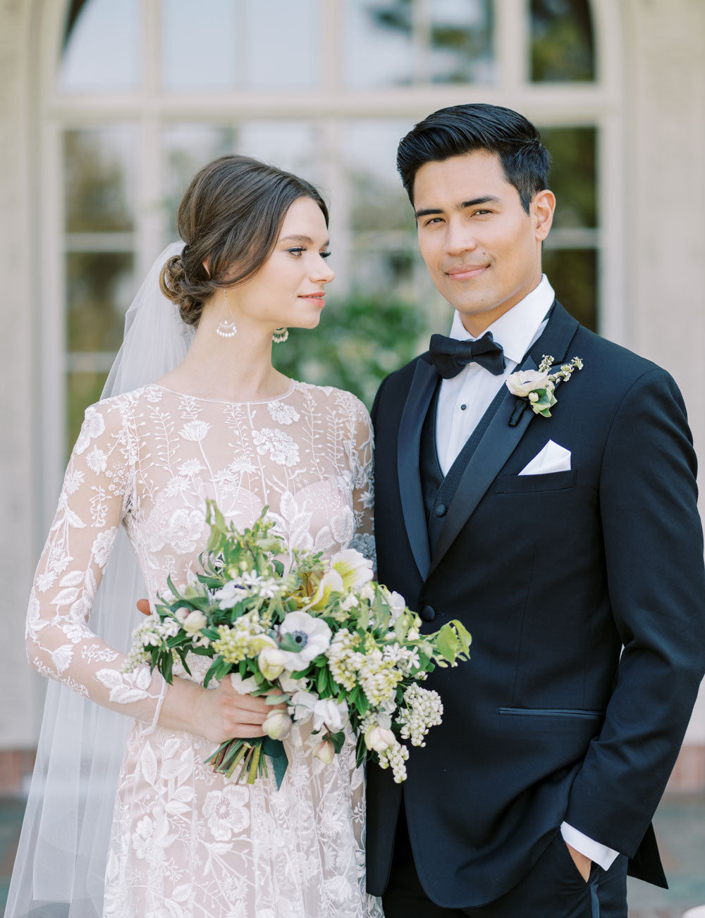 Groom wearing black peak lapel tuxedo with black satin vest and black satin bowtie standing with bride in garden