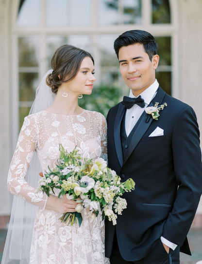 Groom wearing black peak lapel tuxedo with black satin vest and black satin bowtie standing with bride in garden