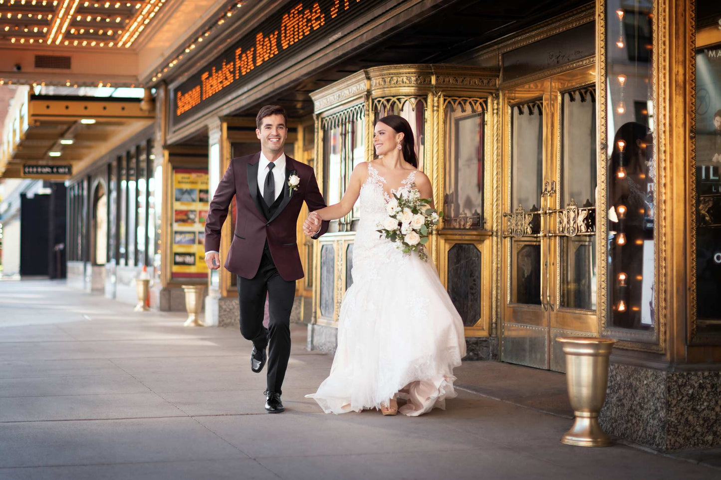 Man in burgundy tuxedo and woman in a wedding gown walking in front of a theater entrance.