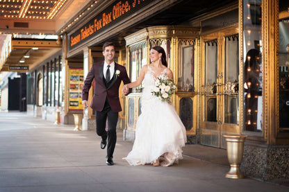 Man in burgundy tuxedo and woman in a wedding gown walking in front of a theater entrance.