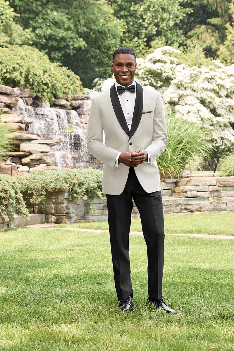 Man in a ivory tuxedo with black lapels, while standing in a beautiful garden with a waterfall on a summer day.