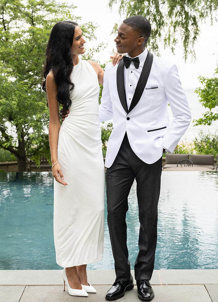 Handsome black man wearing a white tuxedo with black lapel from tuxclub standing with a pretty lady in front of a pool with trees.