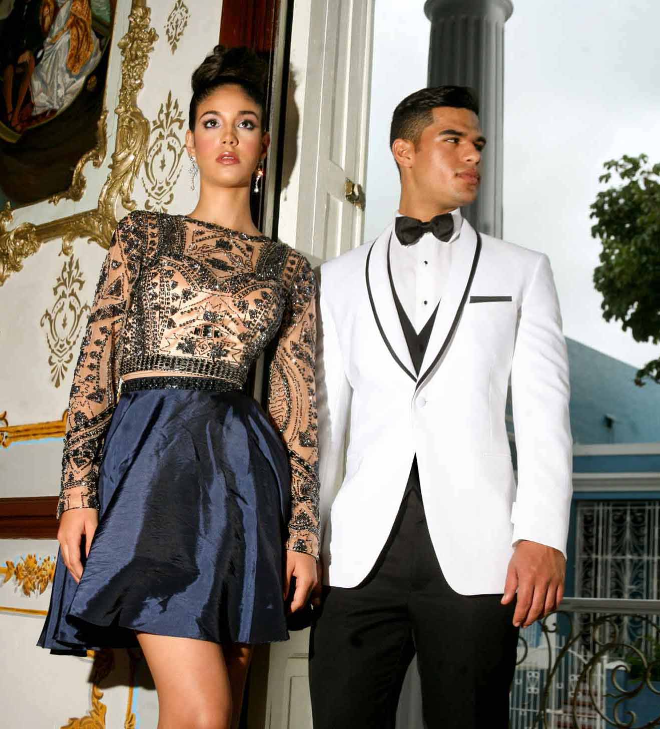 Man in a white shawl collar tuxedo and woman in a black and gold dress standing in an elegant interior setting.