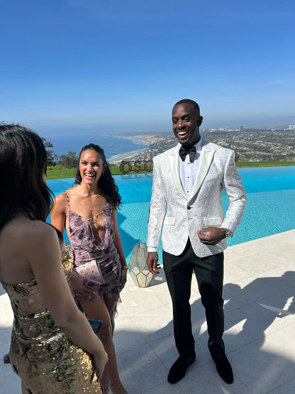 handsome man in a white peak lapel tuxedo with silver highlights and a black bowtie standing next to a pool with a scenic background