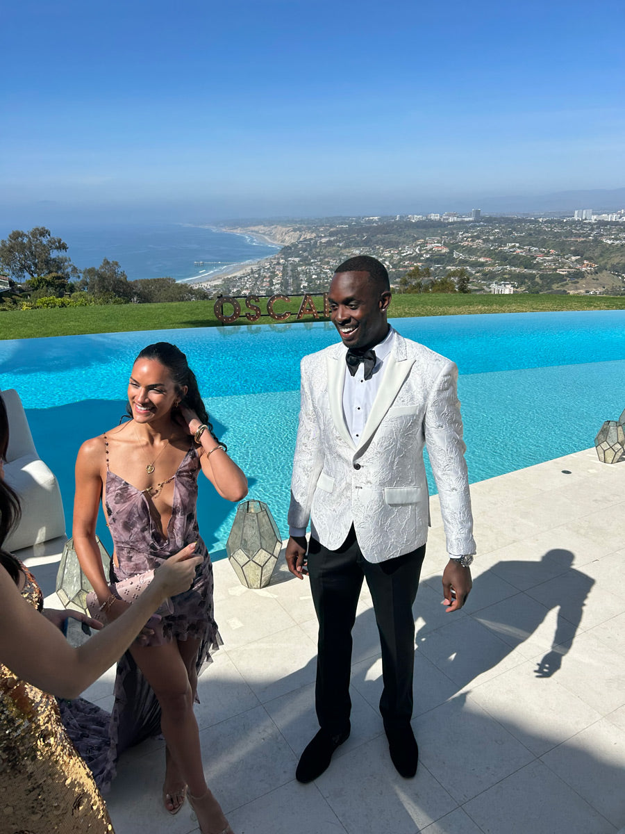 Man in a white peak lapel tuxedo with silver highlights standing next to a pool with a beautiful view in the background